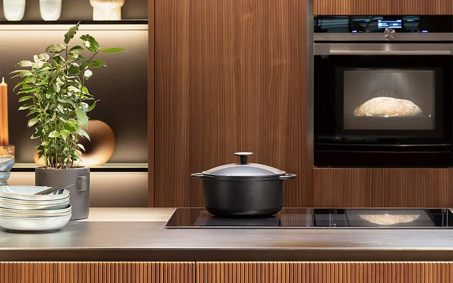 Eine moderne Küche mit einem Induktionsherd und einem schwarzen Topf darauf, hölzernen Groneck-Schränken, einem in die Schränke eingebauten Mikrowellenherd und einer Topfpflanze neben einem Stapel