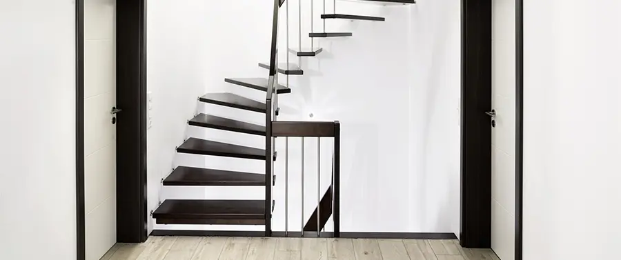 Ein moderner Treppenbau mit dunklen Holzstufen und einem Metallgeländer, vor einem weißen Hintergrund mit einer Glasabtrennung.