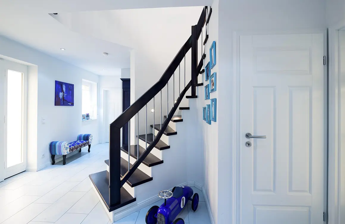 Ein helles, modernes Foyer mit weißen Wänden, einer schwarzen Treppe mit Stufen auf Beton, die in die obere Ebene führt, einer geschlossenen weißen Tür auf der rechten Seite und einem Wohnbereich im Hintergrund mit