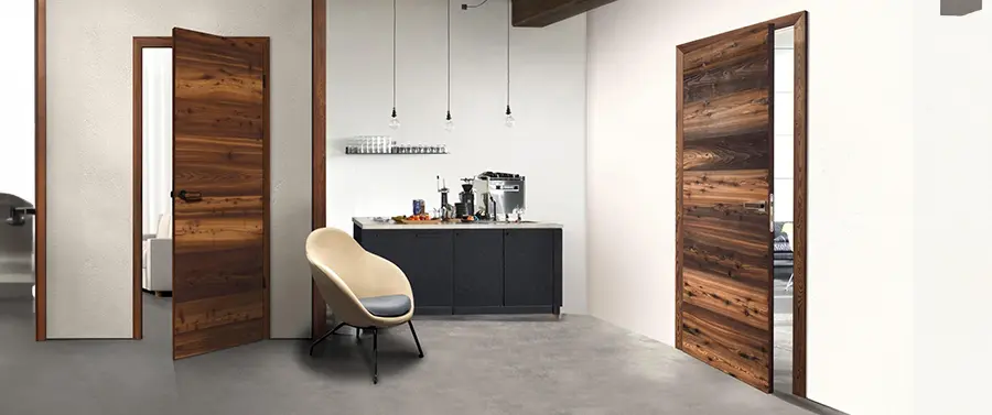 Ein minimalistischer Küchenraum mit einer offenen Holztür mit eleganten Türgriffen, modernen Schränken, einer Kücheninsel mit Kaffeemaschine und verschiedenen Flaschen sowie einem einzelnen Designerstuhl. Das Zimmer verfügt über ein