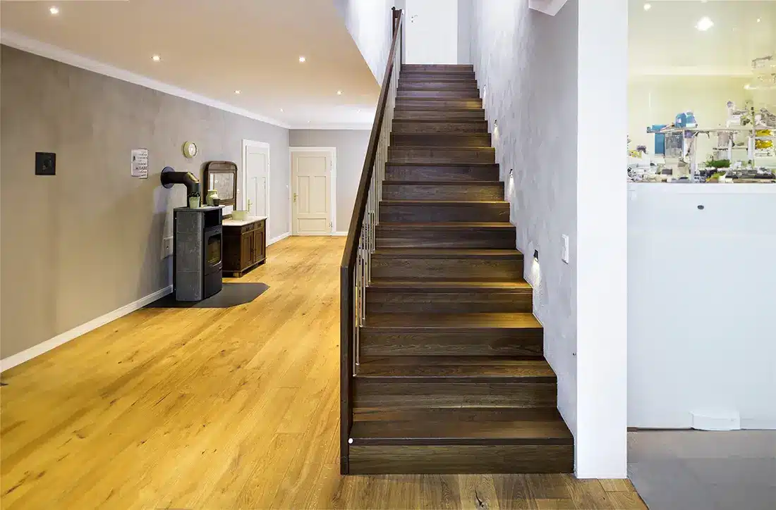 Modernes Interieur eines Wohnhauses mit Faltwerktreppen mit Metallgeländer, die in die obere Ebene führen; Links ist ein Flur mit einer Bank und einem Spiegel,