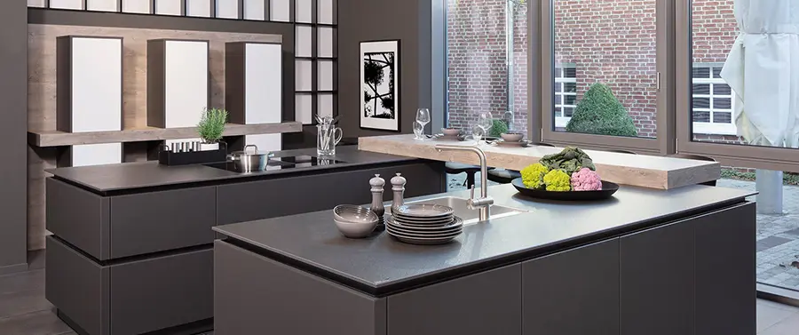 Moderne Kücheneinrichtung mit dunklen Schränken, Spüle aus Edelstahl und einer zentralen Insel mit einer Schüssel Gemüse und einem Tellerstapel, entworfen vom Küchenstudio Melle.