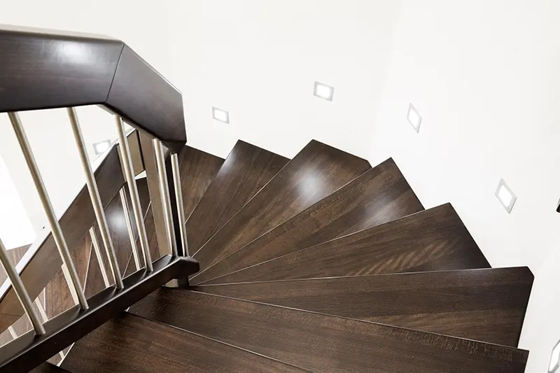 Eine moderne Treppe mit Stufen auf Beton aus dunklem Holz und einem Metallhandlauf führt nach unten vor einem Hintergrund aus weißen Wänden, die mit quadratischen Wandleuchten geschmückt sind.
