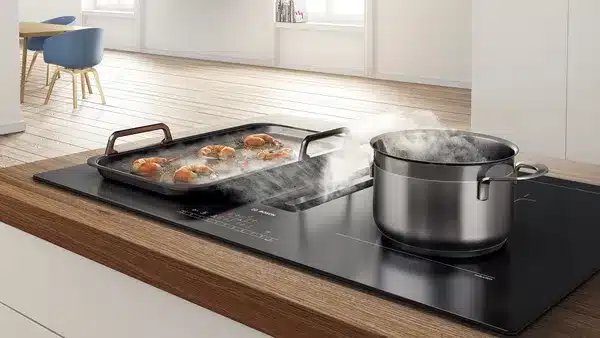 Im Einsatz ist ein modernes Bosch-Induktionskochfeld, auf dessen Oberfläche Dampf aus einem Topf mit brutzelndem Essen aufsteigt und ein silberner Topf darauf steht.