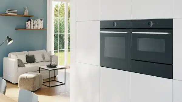 Eine moderne Küche verfügt über eingebaute Doppelöfen von Bosch, eingebettet in elegante weiße Schränke, mit Blick auf ein helles Wohnzimmer mit modernen Möbeln.
