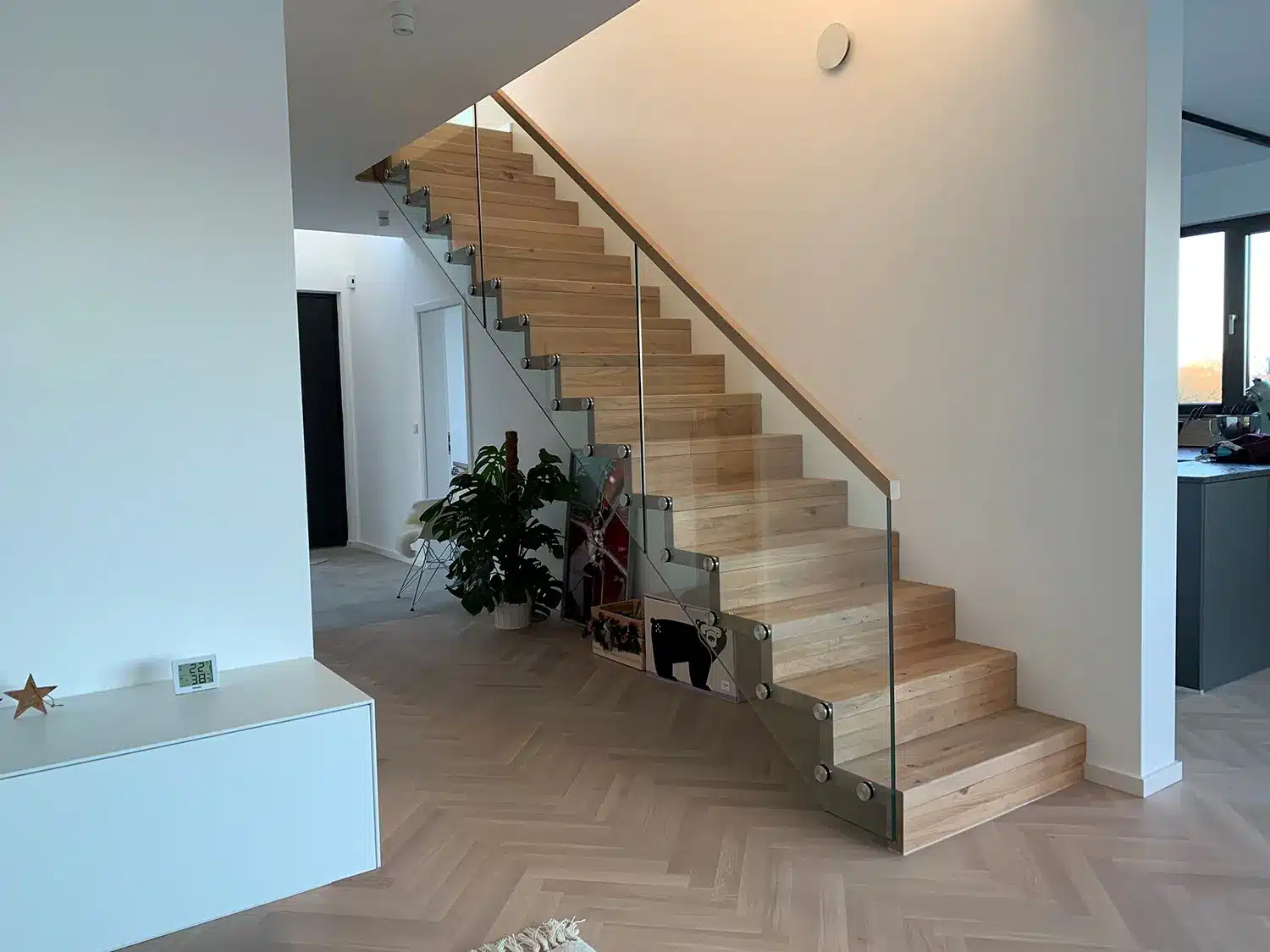Ein moderner Innenraum mit einer Faltwerktreppe mit Glasbalustrade, die in ein Obergeschoss führt, mit Blick auf einen modernen Küchenbereich auf der rechten Seite.