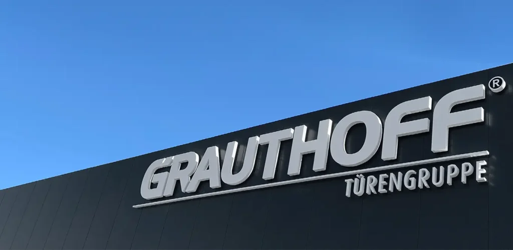 Schild an einer Gebäudefassade mit der Aufschrift „Grauthoff Türen“ vor strahlend blauem Himmel.