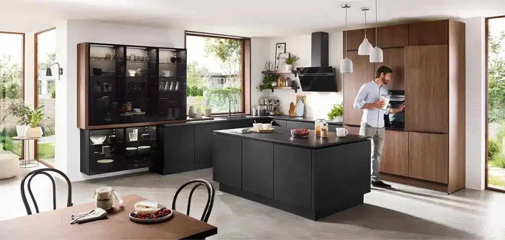 Ein Mann steht in einer modernen Küche mit dunklen Schränken und nippt aus einer Tasse neben einem offenen Kühlschrank, mit einer Kücheninsel im Vordergrund und einem gut beleuchteten Essbereich mit klassischer Küche