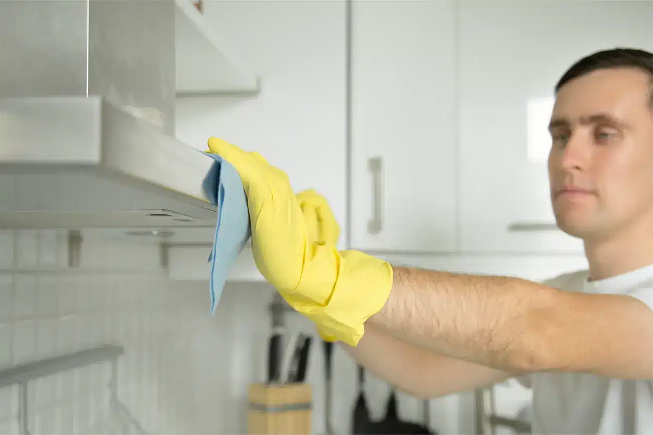 Eine Person mit gelben Handschuhen führt Küchenpflege durch, indem sie eine Dunstabzugshaube mit einem blauen Tuch reinigt.