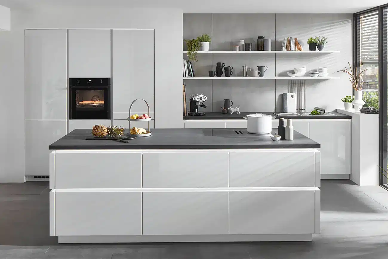 Eine moderne Küche mit eleganten weißen Schränken, einer zentralen Insel, Einbaugeräten und offenen Regalen, dekoriert mit minimalistischen Accessoires und Topfpflanzen. Die Küchenplanung sorgt dafür, dass Funktionalität erfüllt wird