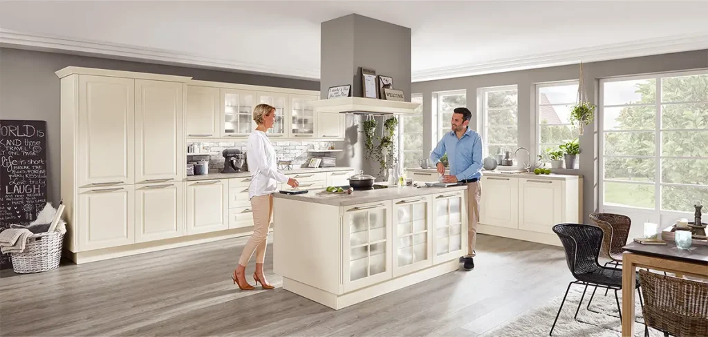 Ein Mann und eine Frau stehen in einer geräumigen, gut beleuchteten Landhausküche mit modernen Geräten und cremefarbenen Schränken. Der Mann interagiert mit der Frau, die daneben steht