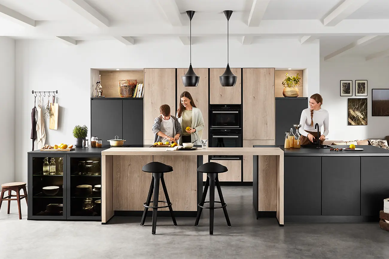 Zwei Personen bereiten in einer modernen Natural Living Küche mit zentraler Kücheninsel Speisen zu, während eine dritte Person an einer Arbeitsplatte sitzt. Die Küche ist mit modernen Schränken und Pendelleuchten ausgestattet.