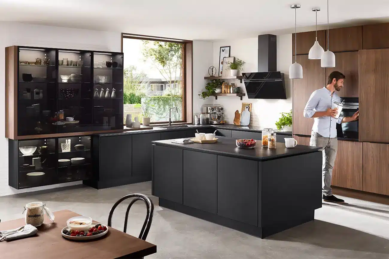 Ein Mann steht in einer modernen Natural Living Küche mit dunklen Arbeitsplatten und blickt in einen offenen Kühlschrank mit einer Kücheninsel im Vordergrund und einer Pendelleuchte darüber.