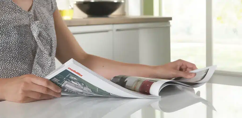 Eine Person sitzt an einem Tisch und blättert in einem Nobilia-Küchenkatalog mit großen Fenstern im Hintergrund.
