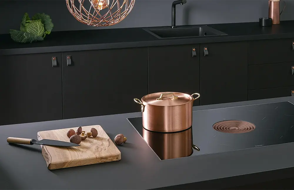 Ein Kupfertopf steht auf einem modernen Bora-Induktionsherd in einer eleganten Küche mit schwarzen Schränken, daneben ein hölzernes Schneidebrett mit einem Messer und verstreuten Haselnüssen.
